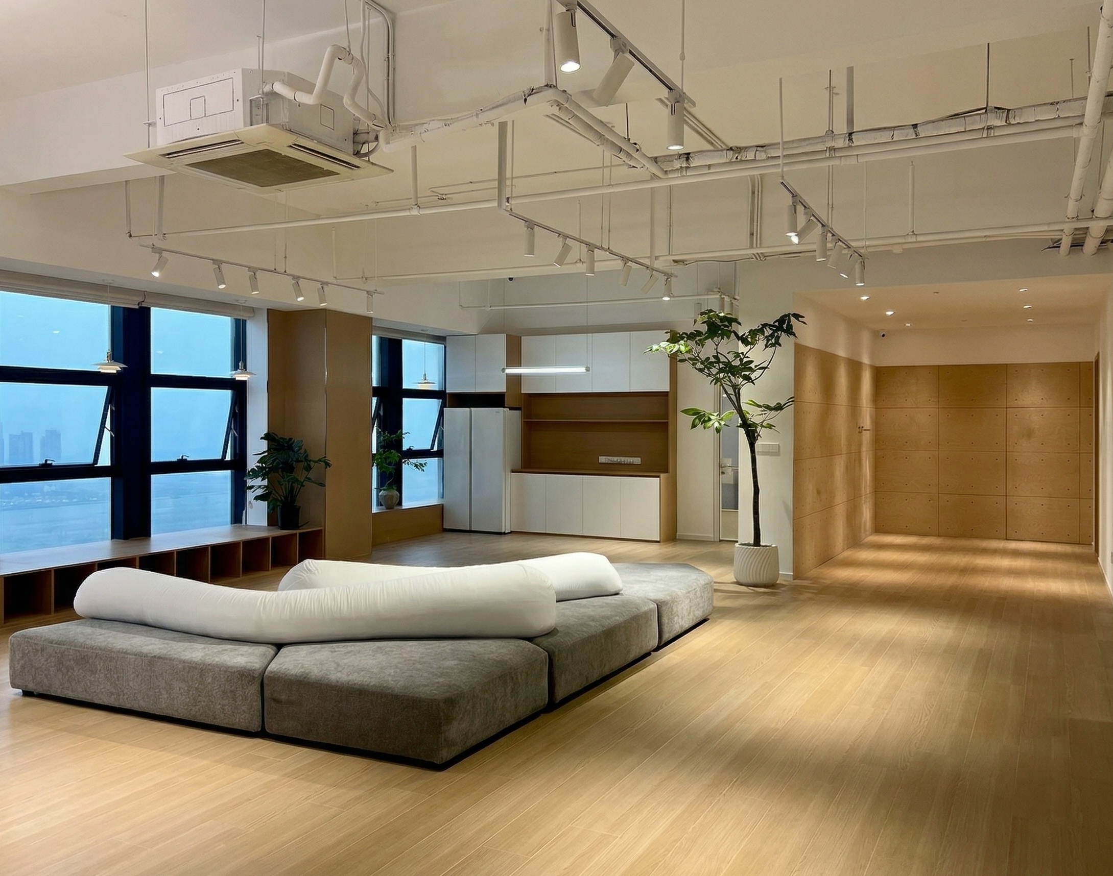 图片展示的是贾维斯联盟·杭州滨江“AI联合战队”场地的公区。画面中,左侧有大窗户,窗外景色隐约可见。窗边摆放着绿植,增添生机。地面铺设木质地板,显得温暖且有质感。左侧还有一排书架,书架旁是灰色的沙发,沙发上铺着白色靠垫。右侧是木质墙面,整体空间明亮且现代,与上下文介绍的“不止是办公室,更是‘AI超级实验室’”相契合,体现了空间的开放与舒适。