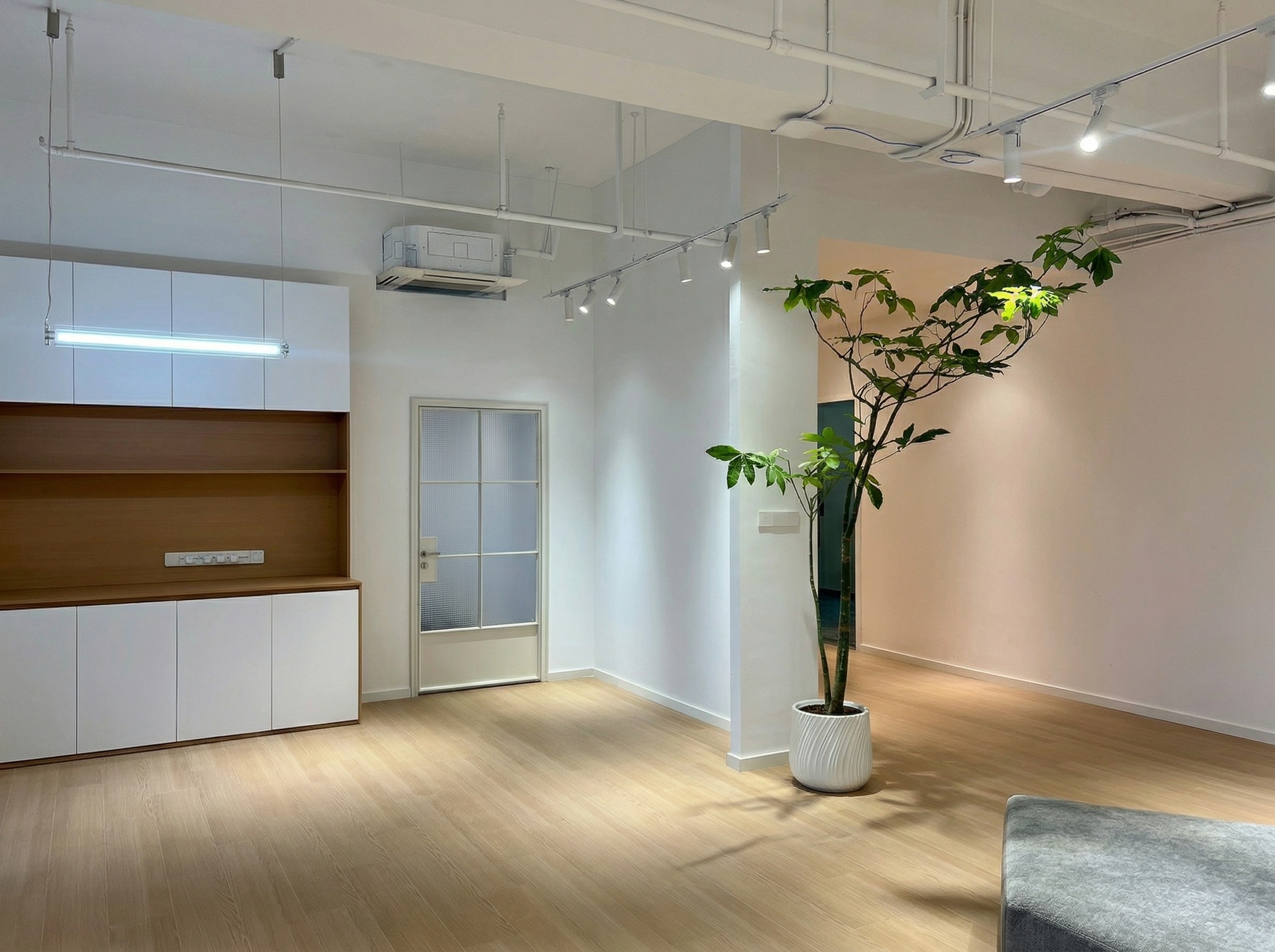 图片展示的是贾维斯联盟·杭州滨江“AI联合战队”场地的实拍场景。画面中,左侧有木质和白色柜体组合的储物区,上方装有空调。右侧是玻璃门,旁边放置着一盆高大的绿植。地面为木质地板,天花板上装有嵌入式灯具。该图片与文档中场地信息部分相关,直观呈现了场地的室内环境,与文档中不止是办公室,更是“AI超级实验室”的描述相呼应。