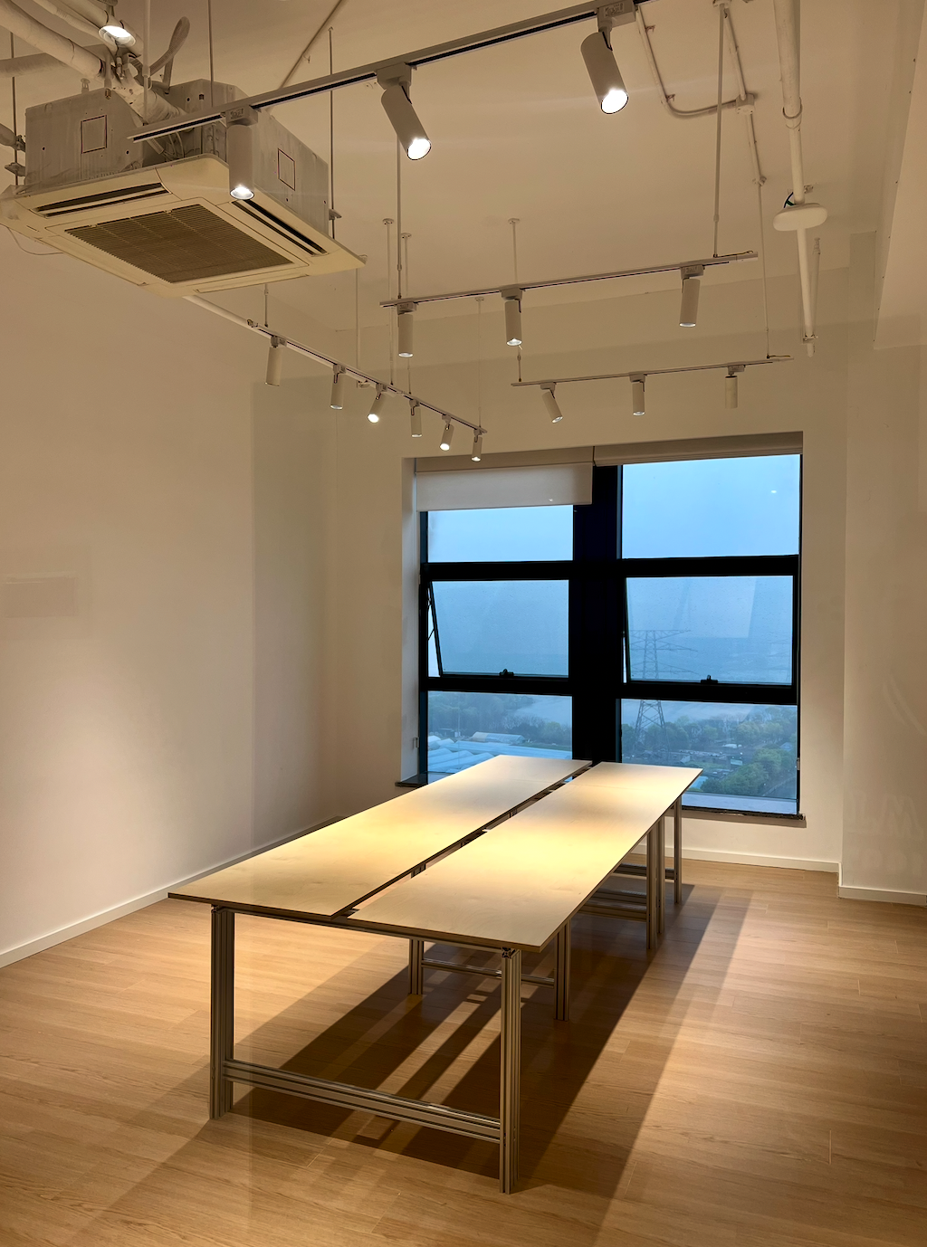 图片展示的是贾维斯联盟·杭州滨江“AI联合战队”场地内的一处公区。画面中,两排长桌并列摆放,桌面呈浅色,桌腿为金属材质。房间内有木质地板,天花板上安装有多个射灯和空调设备。背景是一扇大窗户,窗外景色隐约可见。该图片与文档中介绍场地信息的内容相关,直观呈现了场地内的部分环境。