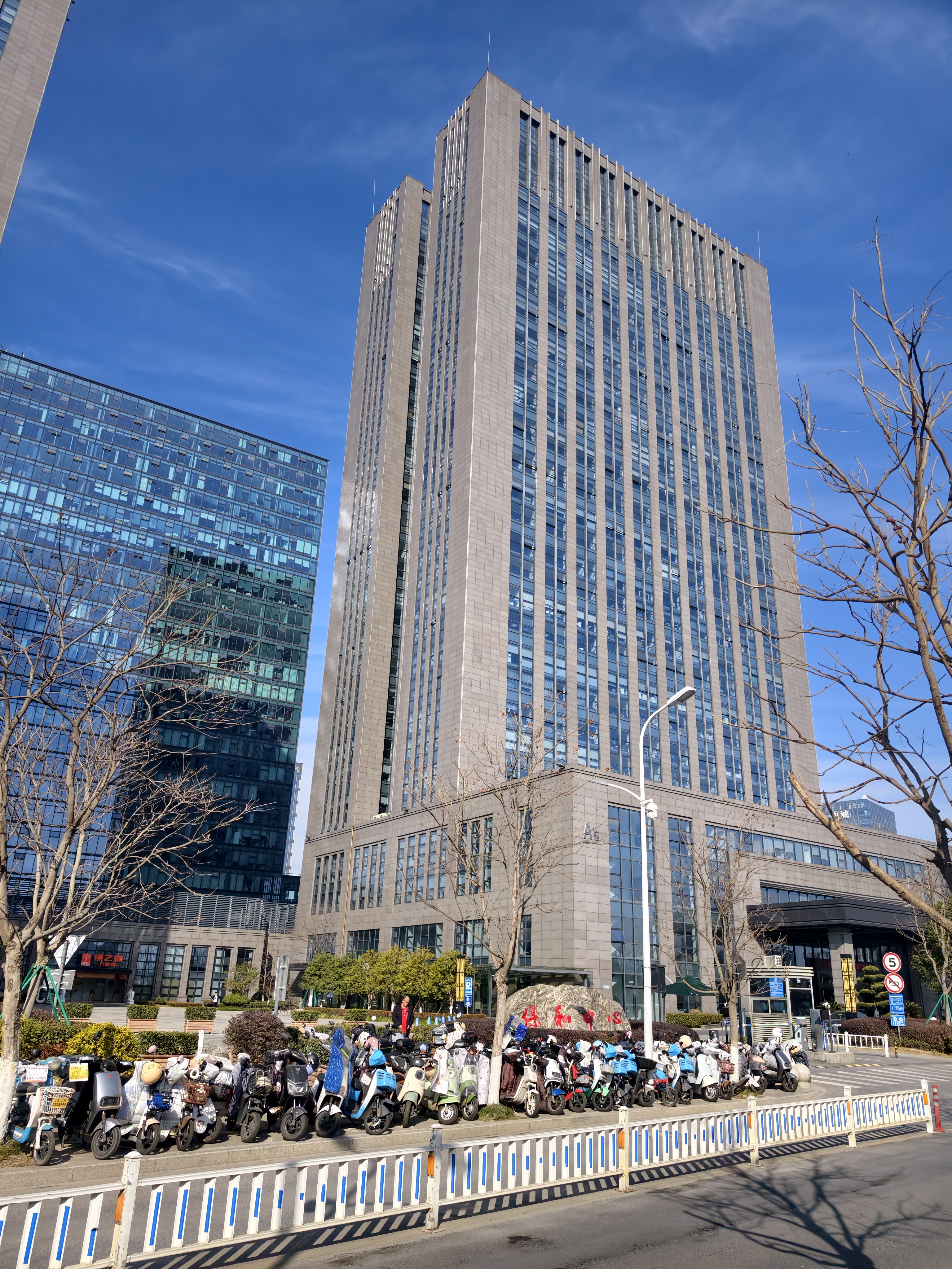 图片展示的是贾维斯联盟·杭州滨江“AI联合战队”场地外观。画面中,一座高耸的现代化玻璃幕墙建筑在蓝天白云下显得格外醒目,其前有整齐停放的电动车。建筑旁有几棵光秃秃的树木,地面上有蓝色和白色相间的护栏。建筑左侧还有一栋玻璃幕墙建筑。图片与文档中场地信息部分对应,直观呈现了场地外观,为读者提供视觉参考。